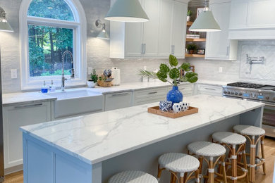 Kitchen - large coastal light wood floor kitchen idea in Charlotte with a farmhouse sink, shaker cabinets, white cabinets, quartzite countertops, white backsplash, marble backsplash, stainless steel appliances, an island and white countertops