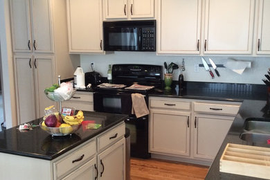 Inspiration for a mid-sized transitional l-shaped light wood floor and brown floor eat-in kitchen remodel in Denver with a double-bowl sink, recessed-panel cabinets, white cabinets, granite countertops, black appliances, an island, black backsplash and black countertops