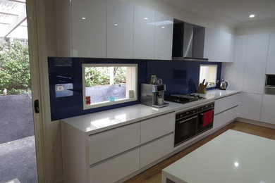This is an example of an expansive modern l-shaped open plan kitchen in Melbourne with a double-bowl sink, white cabinets, quartz benchtops, blue splashback, glass sheet splashback, stainless steel appliances, light hardwood floors and with island.