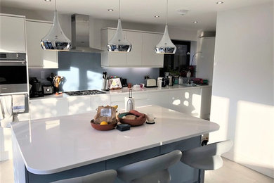 Design ideas for a medium sized contemporary single-wall open plan kitchen in Hertfordshire with an integrated sink, flat-panel cabinets, white cabinets, quartz worktops, blue splashback, glass sheet splashback, stainless steel appliances, porcelain flooring, an island, beige floors and white worktops.