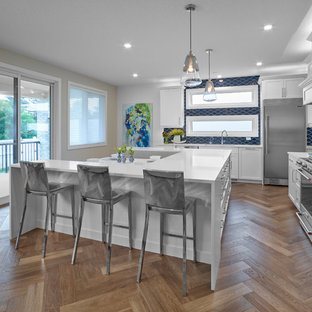 Mid-sized craftsman kitchen pantry designs - Example of a mid-sized arts and crafts u-shaped medium tone wood floor and brown floor kitchen pantry design in Edmonton with a double-bowl sink, shaker cabinets, white cabinets, quartz countertops, blue backsplash, glass tile backsplash, stainless steel appliances, an island and white countertops