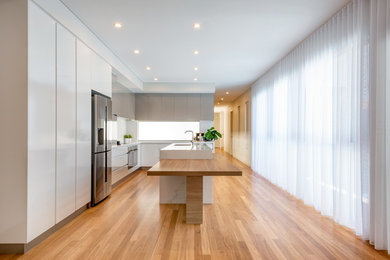 This is an example of a large galley open plan kitchen in Perth with a drop-in sink, flat-panel cabinets, white cabinets, wood benchtops, brown splashback, ceramic splashback, stainless steel appliances, medium hardwood floors, with island, brown floor and white benchtop.