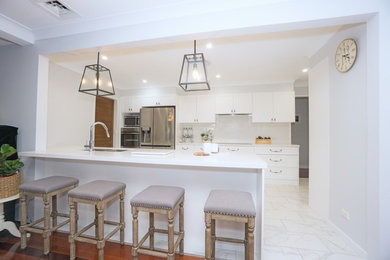 Mid-sized traditional single-wall eat-in kitchen in Sydney with a double-bowl sink, shaker cabinets, white cabinets, quartz benchtops, white splashback, stainless steel appliances, a peninsula, white floor, white benchtop and subway tile splashback.