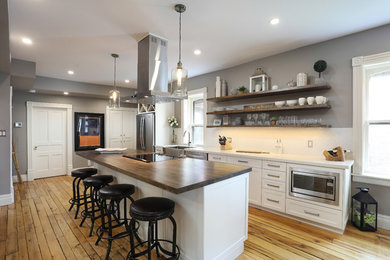 Mid-sized transitional single-wall medium tone wood floor and brown floor eat-in kitchen photo in Other with a farmhouse sink, shaker cabinets, white cabinets, white backsplash, stainless steel appliances, an island and white countertops