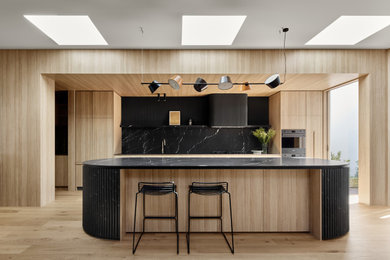 Design ideas for a mid-sized contemporary open plan kitchen in Melbourne with a single-bowl sink, flat-panel cabinets, light wood cabinets, marble benchtops, black splashback, stone slab splashback, black appliances, medium hardwood floors, with island and black benchtop.