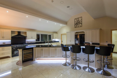 Expansive traditional l-shaped open plan kitchen in Belfast with a belfast sink, beaded cabinets, white cabinets, granite worktops, black splashback, stone slab splashback, black appliances, porcelain flooring, multiple islands, white floors and black worktops.