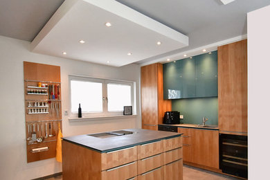 Photo of a large contemporary galley open plan kitchen in Frankfurt with a drop-in sink, flat-panel cabinets, light wood cabinets, solid surface benchtops, green splashback, timber splashback, black appliances, concrete floors, with island, grey floor and grey benchtop.