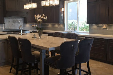 Mid-sized transitional l-shaped travertine floor and beige floor enclosed kitchen photo in Orange County with a farmhouse sink, brown cabinets, marble countertops, gray backsplash, glass tile backsplash, stainless steel appliances, an island and beaded inset cabinets
