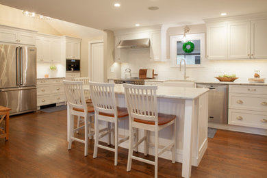 Example of a mid-sized transitional l-shaped medium tone wood floor and brown floor eat-in kitchen design in Bridgeport with a farmhouse sink, recessed-panel cabinets, white cabinets, granite countertops, white backsplash, ceramic backsplash, stainless steel appliances, an island and blue countertops