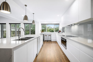 This is an example of a large contemporary l-shaped eat-in kitchen in Sunshine Coast with an undermount sink, flat-panel cabinets, white cabinets, quartz benchtops, grey splashback, glass sheet splashback, stainless steel appliances, dark hardwood floors, with island, brown floor and grey benchtop.