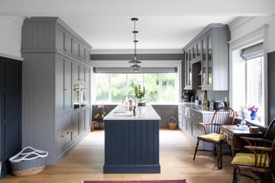 This is an example of a mid-sized country galley open plan kitchen in Other with a farmhouse sink, shaker cabinets, blue cabinets, quartzite benchtops, timber splashback, panelled appliances, light hardwood floors, with island and white benchtop.