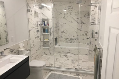 This is an example of a mid-sized contemporary master bathroom in New York with a drop-in tub, a shower/bathtub combo, a one-piece toilet, white tile, porcelain tile, porcelain floors, a drop-in sink, flat-panel cabinets, black cabinets, white walls and solid surface benchtops.