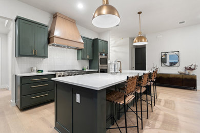 Example of a large l-shaped light wood floor open concept kitchen design in Austin with a farmhouse sink, shaker cabinets, green cabinets, quartz countertops, white backsplash, ceramic backsplash, stainless steel appliances, an island and white countertops