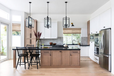 Inspiration for a mid-sized transitional l-shaped kitchen in San Diego with a farmhouse sink, shaker cabinets, granite benchtops, terra-cotta splashback, stainless steel appliances, with island, brown floor, black benchtop, medium wood cabinets, grey splashback and medium hardwood floors.