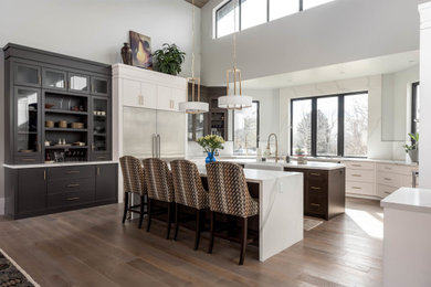 This is an example of a transitional u-shaped kitchen in Salt Lake City with a farmhouse sink, flat-panel cabinets, white cabinets, stainless steel appliances, medium hardwood floors, multiple islands, brown floor and white benchtop.