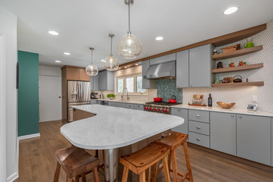 Example of a large mid-century modern l-shaped medium tone wood floor and brown floor kitchen design in Milwaukee with an undermount sink, gray cabinets, quartz countertops, white backsplash, porcelain backsplash, stainless steel appliances, an island and white countertops
