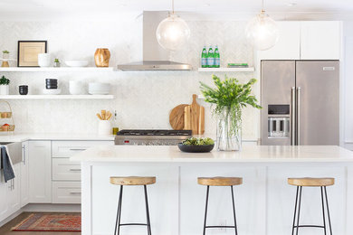 Beach style l-shaped kitchen photo in Miami with a farmhouse sink, shaker cabinets, white cabinets, white backsplash, stainless steel appliances, an island and white countertops