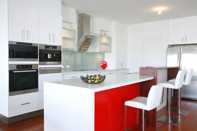 This is an example of a mid-sized contemporary l-shaped eat-in kitchen in Brisbane with an undermount sink, flat-panel cabinets, white cabinets, quartz benchtops, white splashback, glass sheet splashback, stainless steel appliances, medium hardwood floors, with island and brown floor.