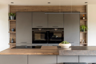 Design ideas for a medium sized contemporary grey and brown single-wall kitchen/diner in Other with a built-in sink, flat-panel cabinets, laminate countertops, brown splashback, black appliances, an island, brown worktops, light hardwood flooring and feature lighting.