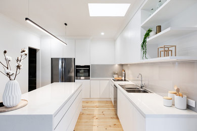Contemporary l-shaped kitchen pantry in Adelaide with a drop-in sink, flat-panel cabinets, white cabinets, quartz benchtops, grey splashback, glass sheet splashback, stainless steel appliances, light hardwood floors, with island, brown floor and white benchtop.