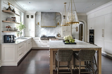 Klassisk inredning av ett stort vit vitt u-kök, med en undermonterad diskho, vita skåp, marmorbänkskiva, stänkskydd i marmor, rostfria vitvaror, mörkt trägolv, en köksö, brunt golv, luckor med infälld panel och grått stänkskydd