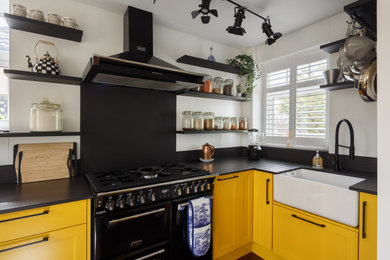 This is an example of a medium sized traditional l-shaped kitchen/diner in Other with a belfast sink, shaker cabinets, yellow cabinets, composite countertops, black splashback, ceramic splashback, black appliances, laminate floors, no island, brown floors, black worktops and a feature wall.