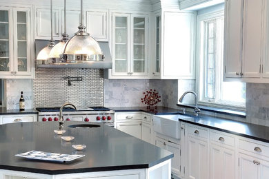 Elegant l-shaped kitchen photo in New York with a farmhouse sink, white cabinets, soapstone countertops, stainless steel appliances, an island, glass-front cabinets, metallic backsplash and metal backsplash