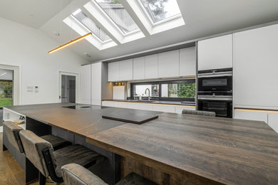 This is an example of a medium sized contemporary grey and brown galley kitchen/diner in Berkshire with a submerged sink, flat-panel cabinets, grey cabinets, granite worktops, black appliances, medium hardwood flooring, an island, brown floors, brown worktops, a vaulted ceiling and feature lighting.