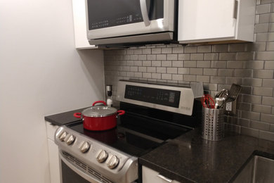 Photo of a small modern galley separate kitchen in Birmingham with a farmhouse sink, flat-panel cabinets, white cabinets, granite benchtops, subway tile splashback, stainless steel appliances, light hardwood floors, no island, beige floor and black benchtop.