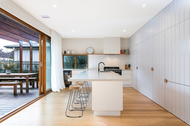 Design ideas for a beach style l-shaped kitchen in Melbourne with an undermount sink, flat-panel cabinets, white cabinets, white splashback, black appliances, medium hardwood floors, with island, brown floor and white benchtop.
