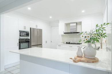 Large transitional u-shaped eat-in kitchen in Sydney with a drop-in sink, shaker cabinets, white cabinets, white splashback, stone slab splashback, stainless steel appliances, no island, grey floor and white benchtop.