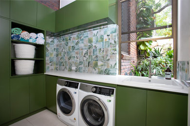 This is an example of a small modern l-shaped laundry room in Brisbane with a drop-in sink, green cabinets, quartz benchtops, green splashback, porcelain splashback, multi-coloured walls, porcelain floors, a side-by-side washer and dryer, grey floor and white benchtop.