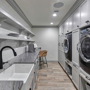 デトロイトの大きいトランジショナルスタイルのおしゃれな洗濯室 (エプロンフロントシンク、落し込みパネル扉のキャビネット、グレーのキャビネット、白い壁、淡色無垢フローリング、ベージュの床、グレーのキッチンカウンター、御影石カウンター) の写真