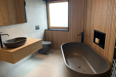 Diseño de cuarto de baño flotante de tamaño medio con puertas de armario de madera clara, bañera exenta, baldosas y/o azulejos marrones, imitación madera, paredes marrones, suelo gris y madera