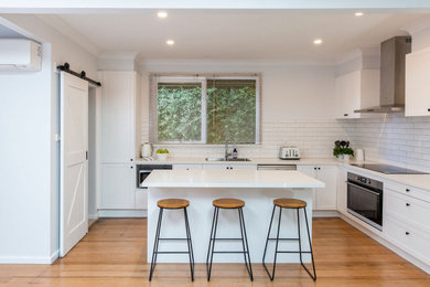Inspiration for a mid-sized modern u-shaped kitchen pantry in Melbourne with a drop-in sink, shaker cabinets, white cabinets, solid surface benchtops, subway tile splashback, stainless steel appliances, medium hardwood floors, with island and white benchtop.