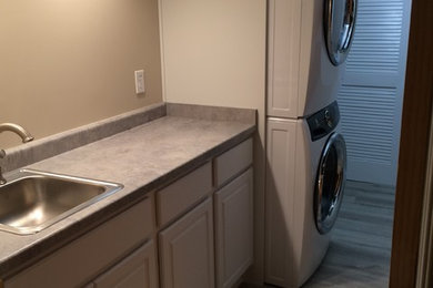 Example of a large trendy galley medium tone wood floor and gray floor dedicated laundry room design in Other with a drop-in sink, raised-panel cabinets, white cabinets, beige walls and a stacked washer/dryer