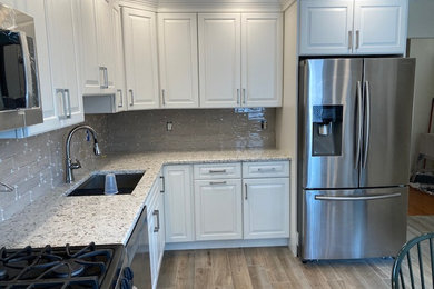 Example of a large minimalist l-shaped light wood floor and brown floor kitchen pantry design in New York with shaker cabinets, white cabinets, quartz countertops, gray backsplash, subway tile backsplash, stainless steel appliances, white countertops, a drop-in sink and no island