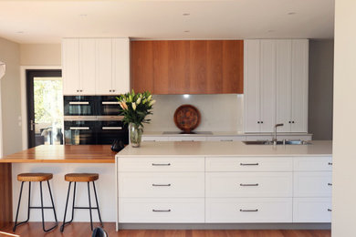 This is an example of a mid-sized contemporary l-shaped eat-in kitchen in Canberra - Queanbeyan with a double-bowl sink, shaker cabinets, white cabinets, quartz benchtops, white splashback, stone slab splashback, black appliances, dark hardwood floors, with island, multi-coloured floor and white benchtop.