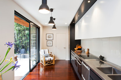 Photo of a mid-sized contemporary single-wall separate kitchen in Sydney with an integrated sink, flat-panel cabinets, stainless steel benchtops, no island, white splashback, glass sheet splashback, stainless steel appliances, medium hardwood floors and brown floor.