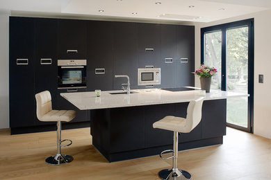 Cette photo montre une cuisine ouverte linéaire tendance de taille moyenne avec un évier intégré, un placard à porte plane, des portes de placard noires, un plan de travail en quartz, une crédence noire, un électroménager en acier inoxydable, parquet clair, îlot et un sol beige.