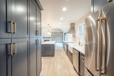 Eat-in kitchen - mid-sized transitional galley medium tone wood floor and gray floor eat-in kitchen idea in New York with a farmhouse sink, shaker cabinets, blue cabinets, quartz countertops, marble backsplash, stainless steel appliances and an island