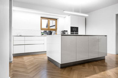 Large contemporary galley eat-in kitchen in Munich with a single-bowl sink, flat-panel cabinets, grey cabinets, solid surface benchtops, white splashback, subway tile splashback, black appliances, light hardwood floors, with island, brown floor and grey benchtop.