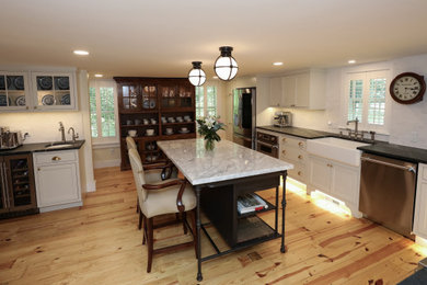 Mid-sized elegant l-shaped light wood floor and brown floor kitchen pantry photo in Boston with a farmhouse sink, beaded inset cabinets, white cabinets, soapstone countertops, white backsplash, marble backsplash, stainless steel appliances, an island and black countertops