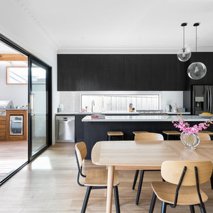 Ejemplo de cocina comedor contemporánea con armarios con paneles lisos, puertas de armario negras, salpicadero de vidrio, electrodomésticos negros, suelo de madera clara, una isla, suelo beige y encimeras blancas