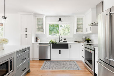 Example of a small transitional l-shaped light wood floor and yellow floor eat-in kitchen design in Vancouver with a farmhouse sink, shaker cabinets, white cabinets, quartz countertops, white backsplash, ceramic backsplash, stainless steel appliances, an island and white countertops