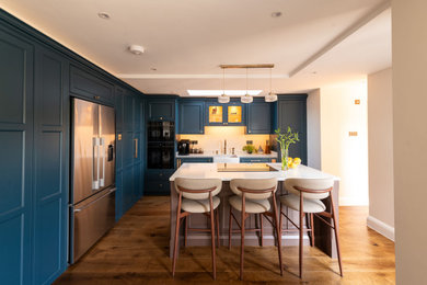 This is an example of a medium sized contemporary l-shaped kitchen/diner in Essex with a belfast sink, shaker cabinets, blue cabinets, engineered stone countertops, white splashback, black appliances, dark hardwood flooring, an island, brown floors, white worktops and a coffered ceiling.