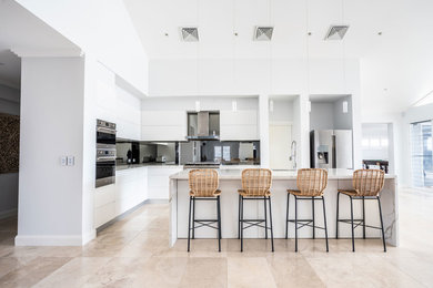 This is an example of a mid-sized contemporary l-shaped open plan kitchen in Sydney with flat-panel cabinets, white cabinets, mirror splashback, white benchtop, an undermount sink, quartz benchtops, metallic splashback, stainless steel appliances, with island and beige floor.