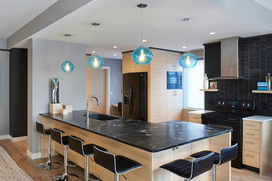 Cette image montre une cuisine ouverte minimaliste en bois clair avec un évier 1 bac, un plan de travail en stéatite, une crédence noire, une crédence en céramique, un électroménager noir, un sol en vinyl et îlot.