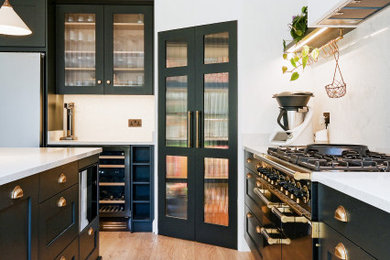 Photo of a large contemporary l-shaped open plan kitchen in London with a belfast sink, shaker cabinets, blue cabinets, granite worktops, white splashback, stone slab splashback, black appliances, light hardwood flooring, an island, beige floors, white worktops, a coffered ceiling and feature lighting.