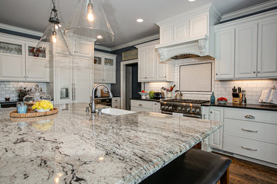 Photo of a large transitional l-shaped open plan kitchen in Charlotte with white cabinets, granite benchtops, white splashback, stainless steel appliances, medium hardwood floors, with island, brown floor, a farmhouse sink, raised-panel cabinets, porcelain splashback and white benchtop.